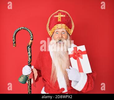 Homme âgé avec cheveux gris et longue barbe costume saint nicholas tenant présent célébrant fou et stupéfié pour succès avec open eyes sc Banque D'Images