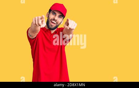 Jeune homme beau avec barbe portant l'uniforme de livraison pointant vers vous et l'appareil photo avec les doigts, souriant positif et gai Banque D'Images