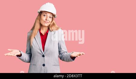 Belle jeune femme portant un architecte hardhat expression sans indice et confuse avec les bras et les mains soulevées. Doute concept. Banque D'Images