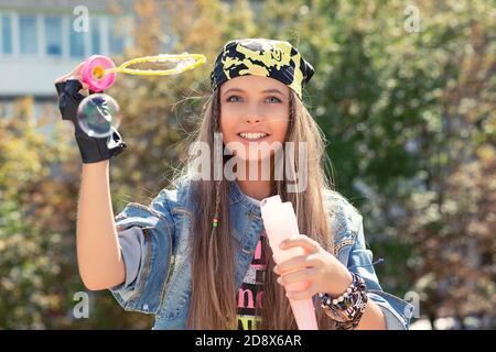 Jeune fille adolescente jeune femme moderne en denim et bandana s'amuser avec des bulles de savon isolé arbres verts ville parc sur fond. Face positive Banque D'Images