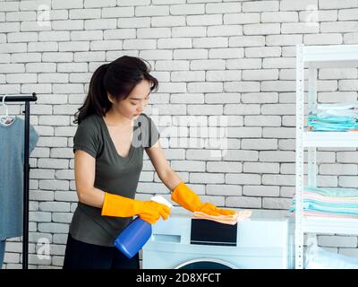 Belle femme asiatique, femme au foyer portant des gants de protection orange en caoutchouc avec un spray nettoyant et un chiffon propre pour nettoyer le lave-linge sur bric blanc Banque D'Images