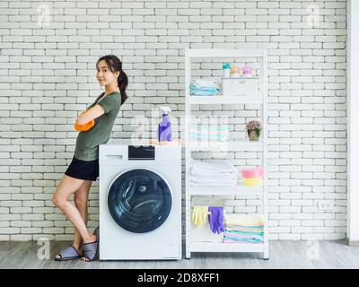 Belle femme asiatique souriante, femme au foyer heureuse portant des gants de protection orange en caoutchouc debout avec les bras pliés à côté de la machine à laver sur brique blanche Banque D'Images
