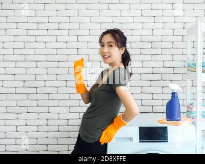 Bonne femme asiatique souriante, femme au foyer portant des gants en caoutchouc orange debout et levant la main dans le geste gagnant et célébrant le succès près de laver ma Banque D'Images