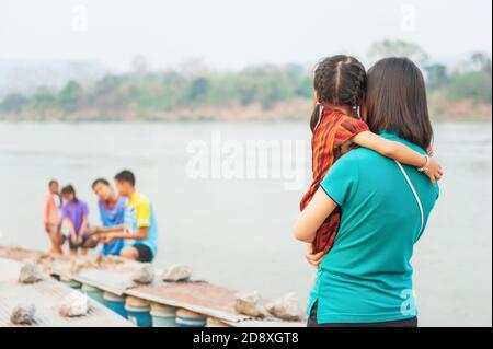 Une petite fille asiatique dans les bras de sa mère se détendant sur la rive dans la soirée. Mékong, Thaïlande. Banque D'Images