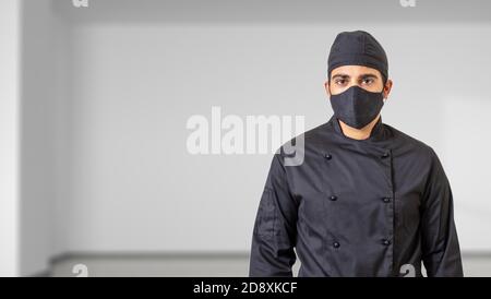 Chef à l'époque des coronavirus. Homme en uniforme de cuisinier avec masque de protection du visage en tissu noir, fond intérieur de la pièce. COVID19 sécurité dans cuisine restaurant f Banque D'Images