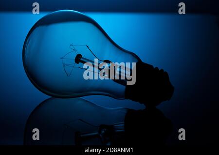 Une ampoule à incandescence repose sur un verre réfléchissant, sur un fond bleu, dans une clé sombre. Banque D'Images