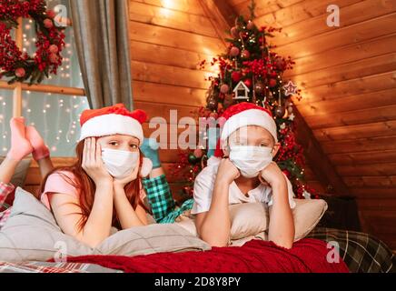 Deux enfants garçon et fille en pyjama, chapeau de père Noël et masques protecteurs sur le lit avoir du plaisir le matin de Noël Banque D'Images