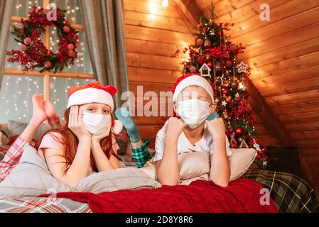 Deux enfants garçon et fille en pyjama, chapeau de père Noël et masques protecteurs sur le lit avoir du plaisir le matin de Noël Banque D'Images