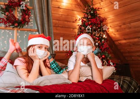 Deux enfants garçon et fille en pyjama, chapeau de père Noël et masques protecteurs sur le lit avoir du plaisir le matin de Noël Banque D'Images
