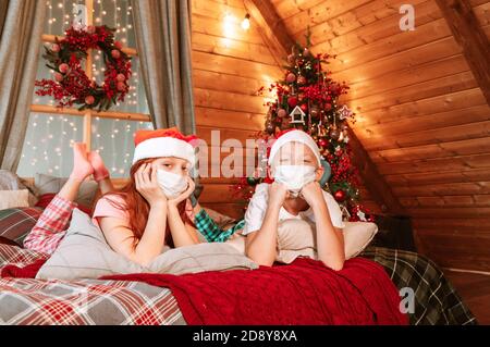 Deux enfants garçon et fille en pyjama, chapeau de père Noël et masques protecteurs sur le lit avoir du plaisir le matin de Noël Banque D'Images