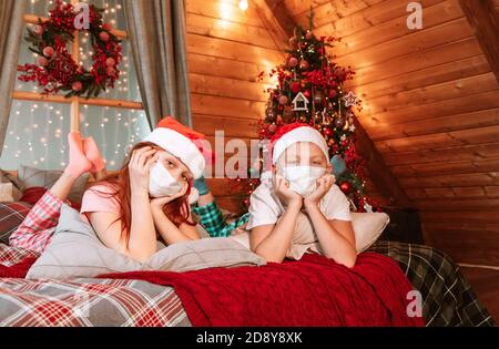 Deux enfants garçon et fille en pyjama, chapeau de père Noël et masques protecteurs sur le lit avoir du plaisir le matin de Noël Banque D'Images