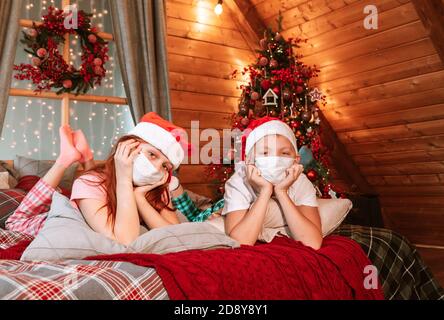 Deux enfants garçon et fille en pyjama, chapeau de père Noël et masques protecteurs sur le lit avoir du plaisir le matin de Noël Banque D'Images
