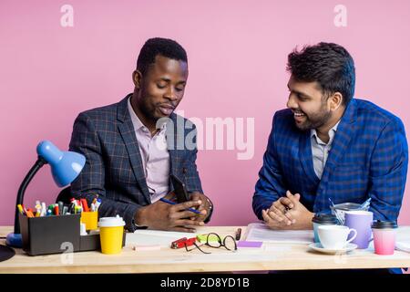 un jeune entrepreneur et un gestionnaire africains discutant d'un projet commun ou des termes de la transaction, tenant des stylos, assis à un bureau au bureau. Entreprise, partenariat, Banque D'Images