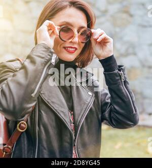 Femme habillée de style boho dans une veste de motard en cuir noir avec sac à rabat en cuir marron ayant une marche de parc d'automne. Banque D'Images