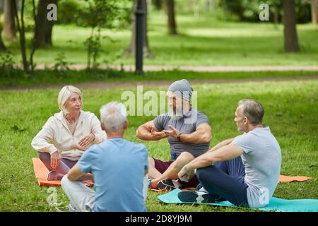 Groupe de personnes âgées actives rassemblées dans le parc de la ville assis sur des tapis à l'écoute de leur entraîneur de yoga Banque D'Images
