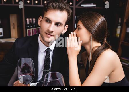 Attention sélective de la femme chuchotant à l'ami en costume près verres de vin au restaurant Banque D'Images