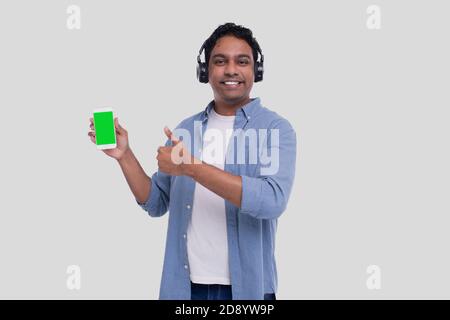 Indian Man écoutant de la musique à partir du téléphone montrant Thoump Up. Homme chantant à la musique. Homme pointant sur l'écran vert du téléphone. Isolé Banque D'Images