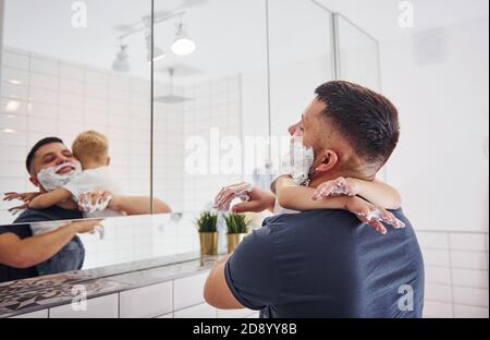 Père avec son fils est dans la salle de bains ont du plaisir en utilisant du gel à raser et en regardant dans le miroir Banque D'Images
