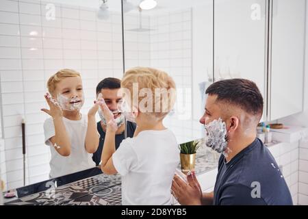 Père avec son fils est dans la salle de bains ont du plaisir en utilisant du gel à raser et en regardant dans le miroir Banque D'Images