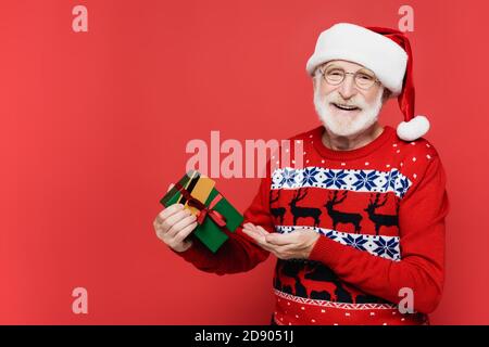 Homme senior souriant dans le chapeau de père noël pointant de la main vers boîte cadeau et carte de crédit isolée sur rouge Banque D'Images