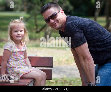 Lors d'un voyage dans le parc, une fille s'assoit sur une table et sourit à la caméra pendant que son père s'accrousse à son niveau Banque D'Images