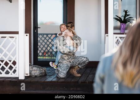 Homme de service militaire souriant embrassant sa fille, assis sur le genou au seuil avec une femme floue au premier plan Banque D'Images