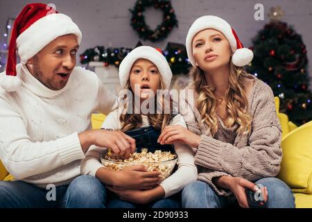 une famille choquée dans des chapeaux de père noël regardant l'appareil photo près du bol avec pop-corn Banque D'Images