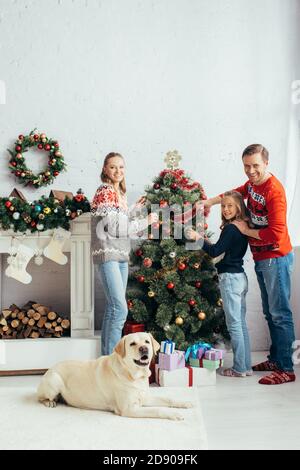 une famille heureuse décorant l'arbre de noël près du labrador dans la salle de séjour Banque D'Images