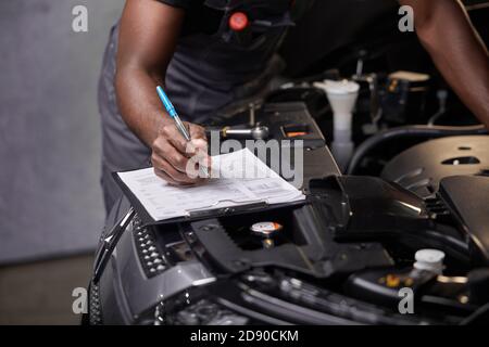 technicien d'entretien automobile professionnel africain en uniforme debout près de la réparation de capot de voiture et en utilisant la liste de contrôle pour l'inspection de voiture, à l'intérieur Banque D'Images