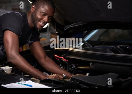 technicien d'entretien automobile professionnel africain en uniforme debout près de la réparation de capot de voiture et en utilisant la liste de contrôle pour l'inspection de voiture, à l'intérieur Banque D'Images