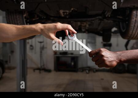 deux jeunes mécaniciens divers mains donnant, partageant l'outil entre eux. artisan automobile pendant le travail, aider, coopérer. concept d'entretien de voiture Banque D'Images