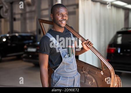 jeune mécanicien automobile africain au travail avec une partie séparée de la machine, posant à la caméra, va réparer Banque D'Images