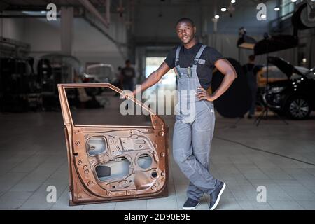 jeune mécanicien automobile africain au travail avec une partie séparée de la machine, posant à la caméra, va réparer Banque D'Images