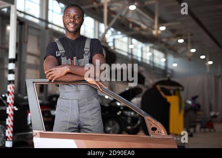 jeune mécanicien automobile africain au travail avec une partie séparée de la machine, posant à la caméra, va réparer Banque D'Images