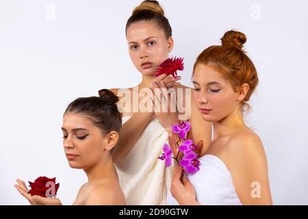 Modèles féminins attrayants avec cheveux tirés vers l'arrière, enveloppés dans des serviettes blanches et tenant des fleurs. Posé sur un fond blanc dans le studio. Banque D'Images