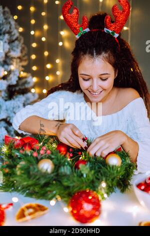 Belle femme souriante portant une robe tricotée blanche faisant couronne de Noël dans la chambre décorée. Banque D'Images