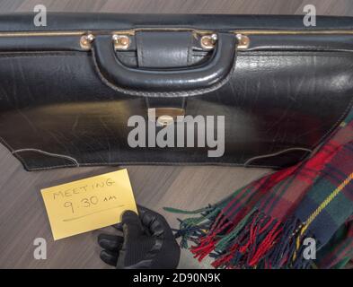 Gros plan de dessus de POV un vieux porte-documents en cuir et une écharpe sur un bureau, avec une main gantée tenant un message confirmant «meeting 9.30 am». Banque D'Images