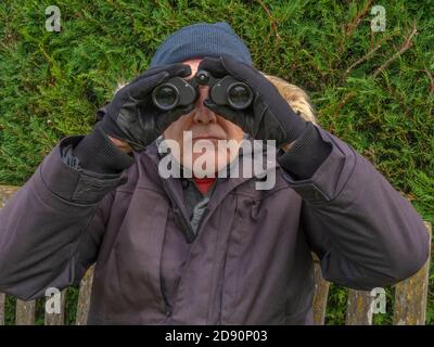 Homme plus âgé vêt d'hiver d'un pardessus, d'un chapeau et de gants, assis sur un banc extérieur, avec une paire de jumelles à ses yeux, regardant vers l'appareil photo. Banque D'Images