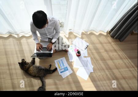 Photo d'un jeune homme travaillant sur un ordinateur portable avec son chat au sol à la maison. Banque D'Images