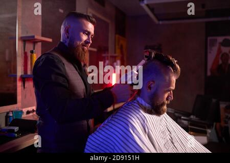 confiant caucasien barber mster coupe les cheveux et la barbe des hommes dans le salon de coiffure, professionnel coiffeur fait la coiffure pour un jeune homme, il travaille avec la tondeuse Banque D'Images