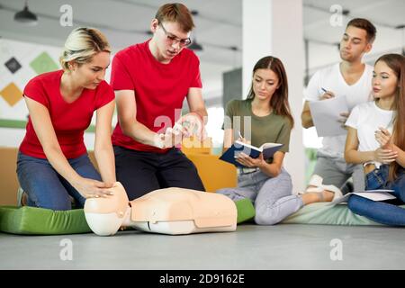 Cours de RCR avec de jeunes instructeurs caucasiens parlant et faisant la démonstration d'aide de premiers soins, mannequin de rcr sur le sol Banque D'Images