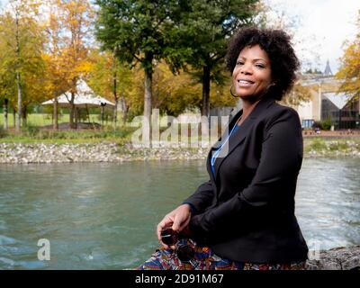 Portrait horizontal d'une femme afro-américaine souriante et attrayante une veste noire et assise près d'une rivière Banque D'Images