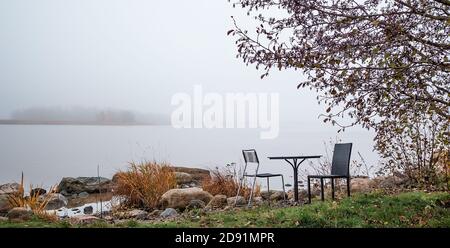 Table et deux chaises sous l'arbre sur la côte en journée de brouillard gris. Paysage de novembre. Un lac avec brouillard blanc dense. Vaporiser sur l'eau. Concept d'automne. Banque D'Images