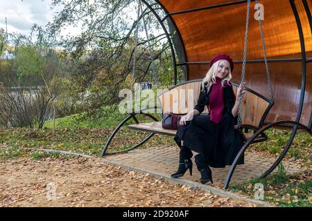 Une femme souriante est assise sur une balançoire de jardin, forgée dans un manteau bordeaux et le biret, avec de beaux yeux, à l'automne sur le fond d'arbres, bleu Banque D'Images