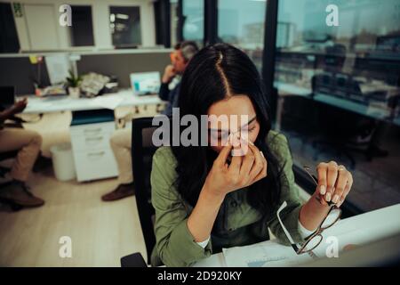Stressé femme d'affaires de race mixte en se grattant ses yeux secs pendant respecter les délais se sentir stressé assis au bureau Banque D'Images