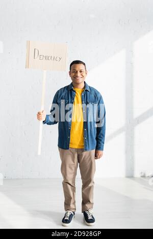 homme indien souriant portant un écriteau avec un lettrage démocratique contre du blanc mur de briques Banque D'Images