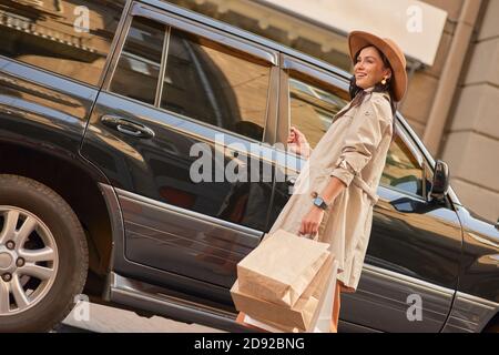 Fin des achats. Jeune femme élégante et heureuse avec ses sacs à provisions en papier, en regardant de côté et en souriant en se tenant contre une voiture de luxe noire dans la rue de la ville. Le mode de vie des gens, la mode Banque D'Images