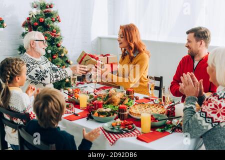 REDHEAD femme saluant homme senior avec boîte cadeau, assis à table avec la famille Banque D'Images