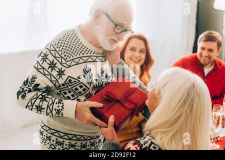 Une femme âgée heureuse prend une boîte cadeau de son mari, assise avec sa famille à la maison Banque D'Images