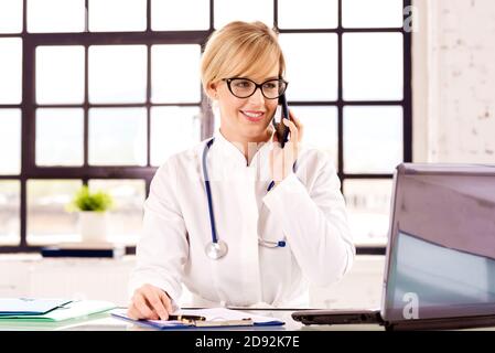 Portrat photo d'une femme souriante médecin assise à son bureau et faisant un appel avec sa patiente tout en travaillant dans le bureau du médecin. Banque D'Images